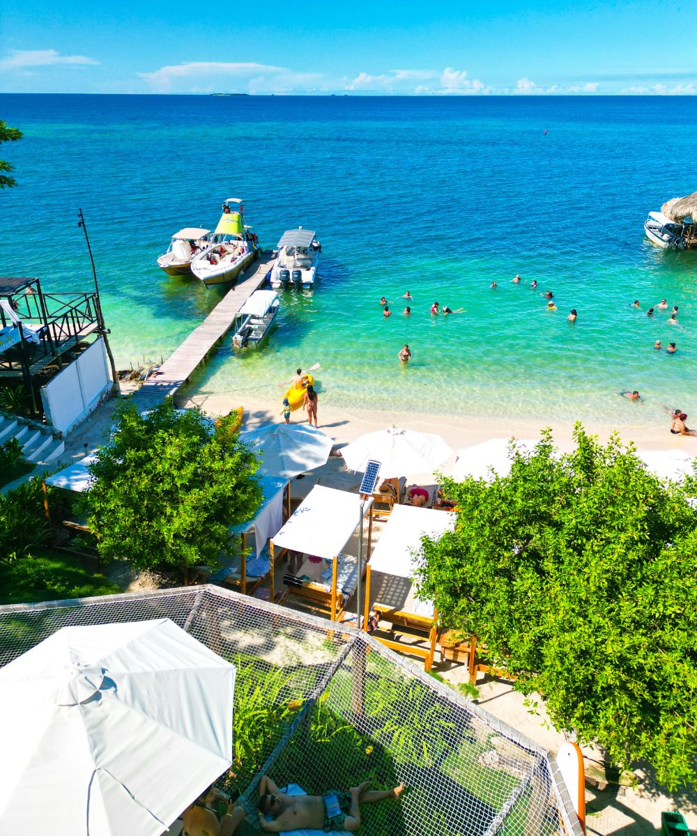Vista de drone de la exclusiva Playa Rosario Beach con camas privadas, asoleadoras blancas y muelle flotante en el mar turquesa.